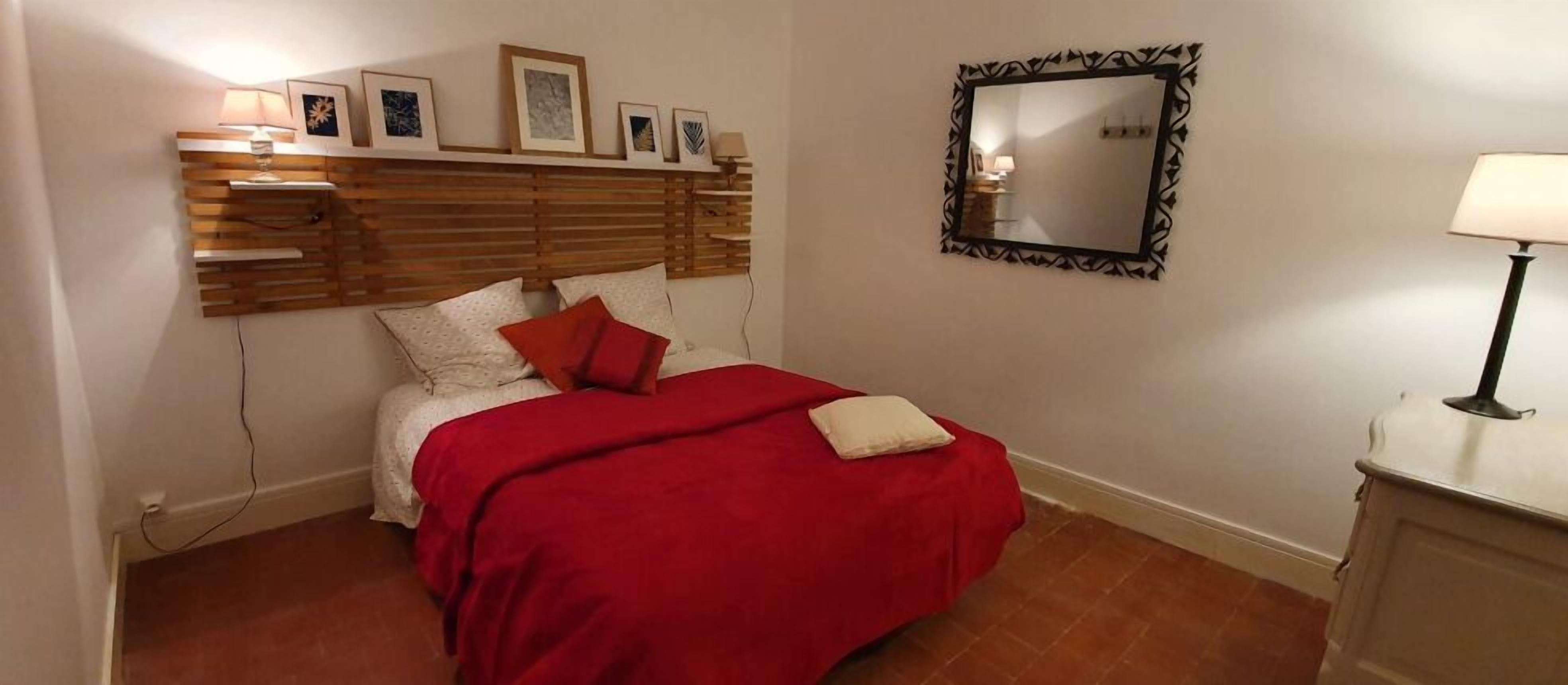 Un dormitorio con cabecera de madera, colcha roja, almohada blanca, espejo y lámpara de noche.