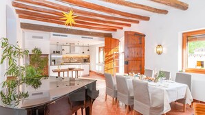 A dining area with a table set for four, a kitchen in the background, and a wooden ceiling with exposed beams.
