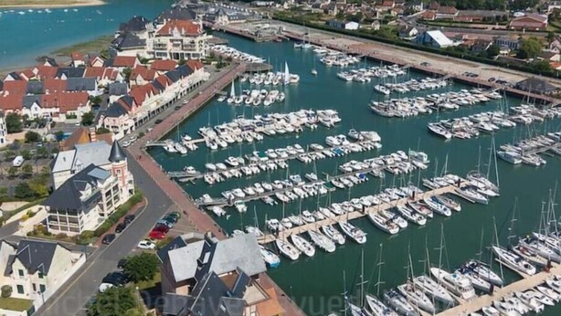 An aerial view of a marina with numerous boats docked, surrounded by residential buildings and a hilly landscape in the background.