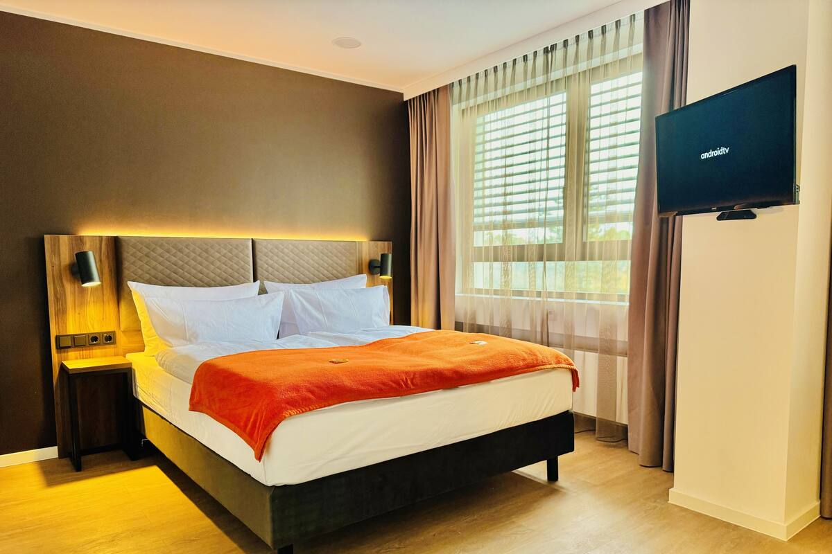 Ein Hotelzimmer mit Bett, Fernseher, Nachttisch und einem Fenster mit Vorhängen.