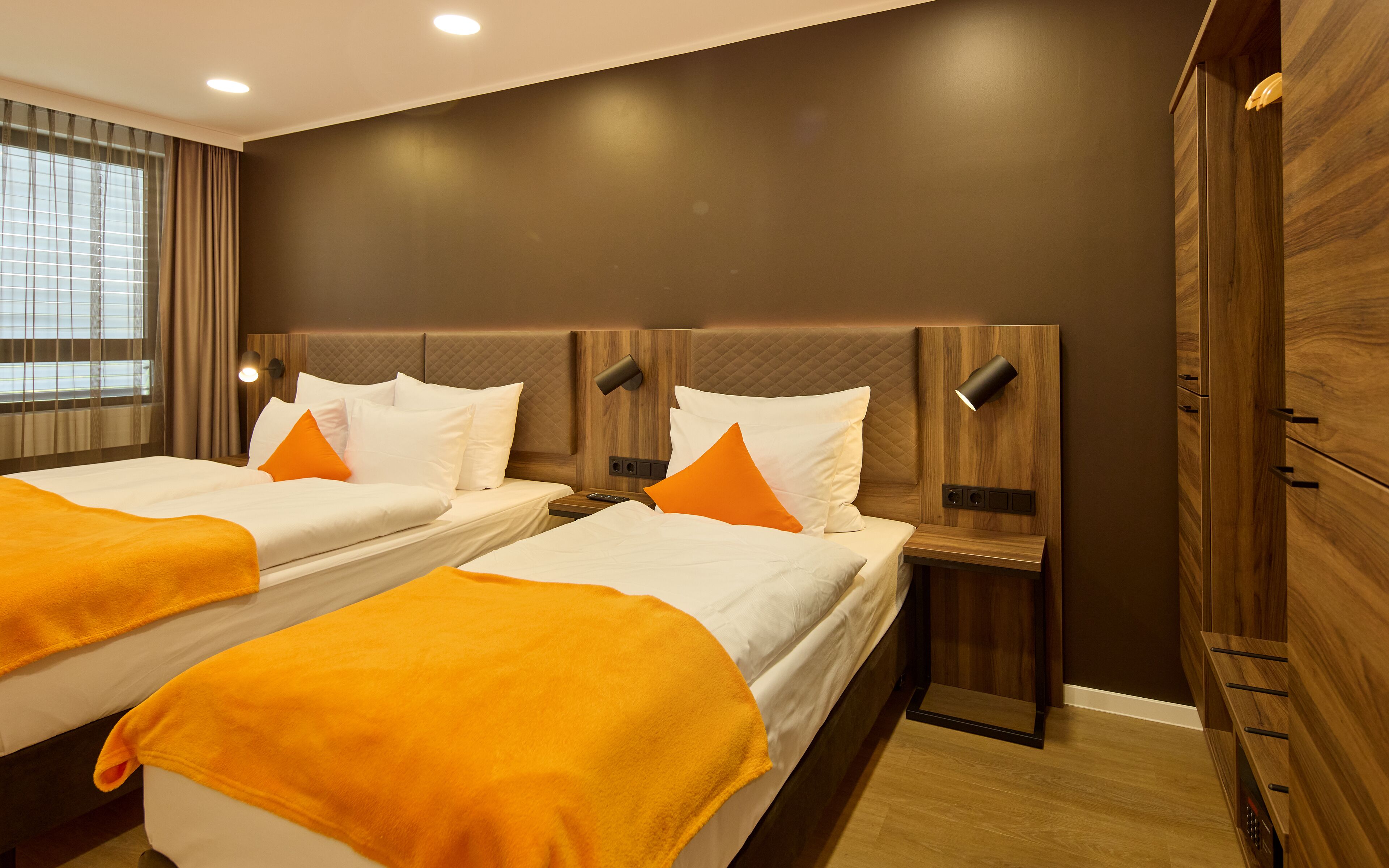 A hotel room with two beds, each with white bedding and orange throw pillows, a wooden headboard, a nightstand with a lamp, and a large wardrobe.