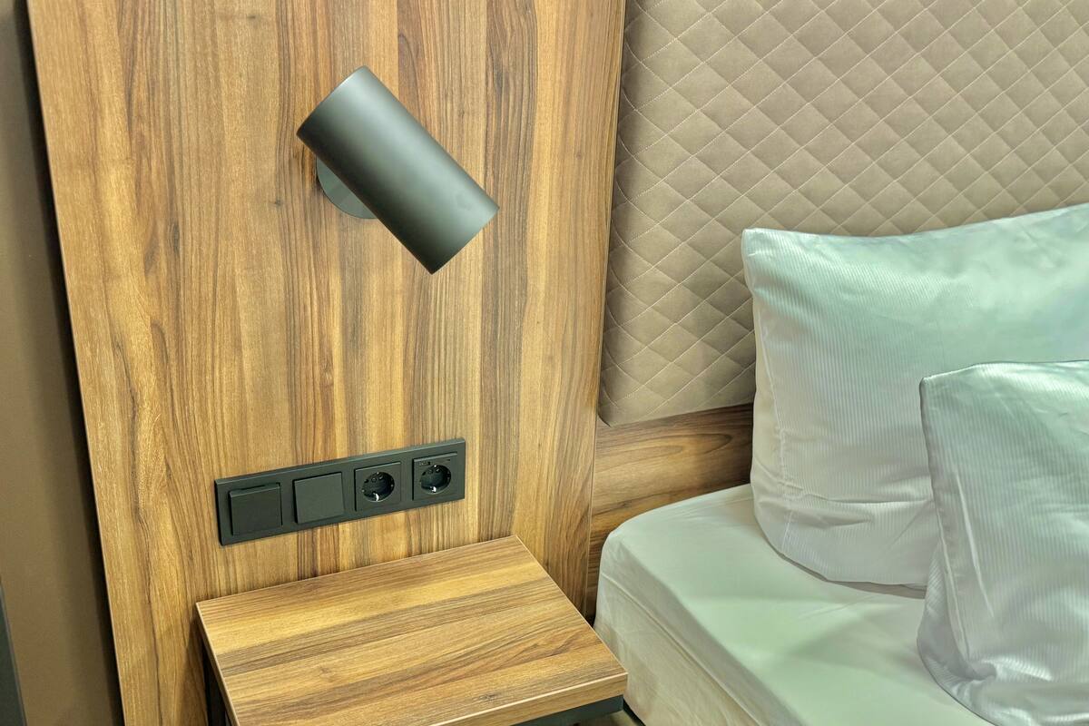 Ein hölzerner Kopfseitenverzierer, ein Lautsprecher in Schwarz, ein Nachttisch und ein Bett mit weißer Bettwäsche.
