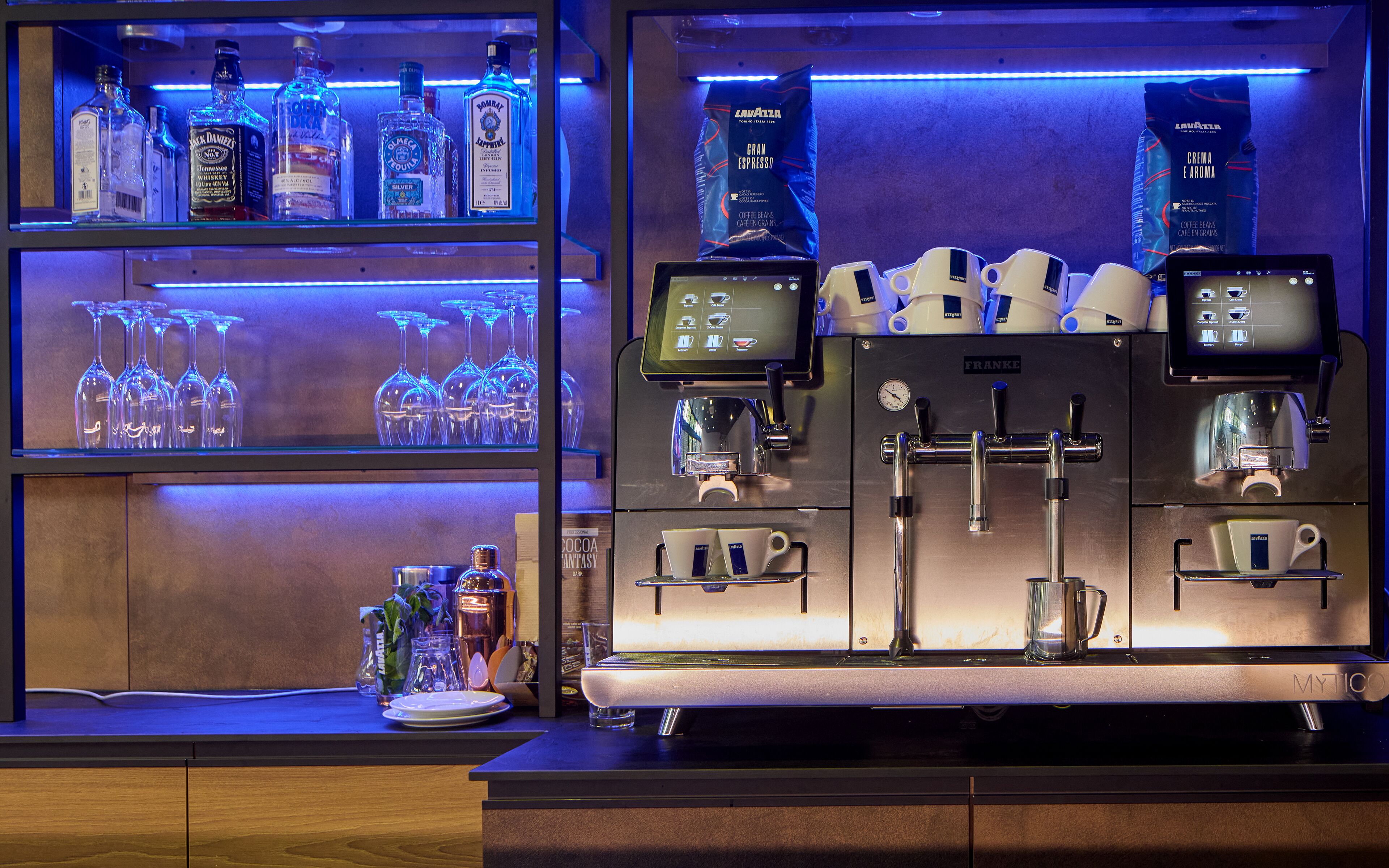 Una cafetera con diversas opciones de preparación y una pantalla, un estante con varias botellas de licor, y una vitrina con copas de vino.
