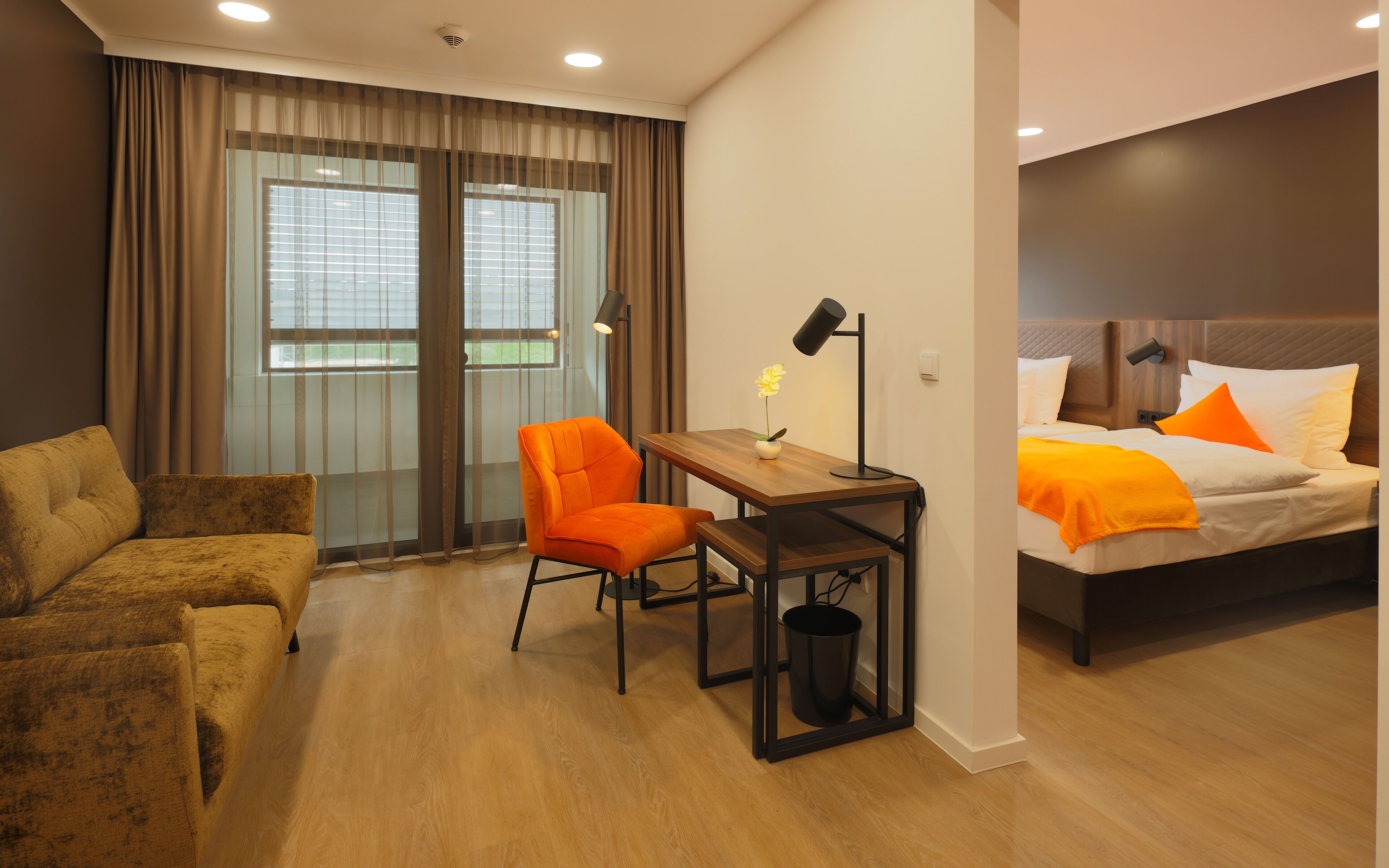 Hotelkamer met een bed, een bureau, een oranje fauteuil, een bank en een raam met gordijnen.