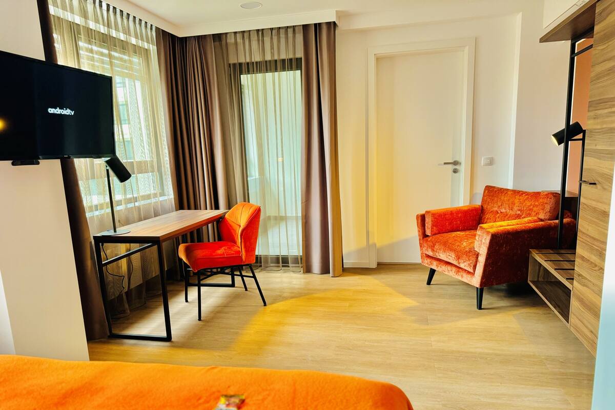 Ein Hotelzimmer mit Holzboden, einem Schreibtisch mit Stuhl, einem Fernseher und einem Bett mit orangefarbener Decke.