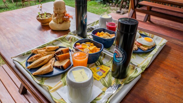A wooden table with a breakfast spread including bread, fruit, and a thermos. - Boas Ondas Pousada (Tibau do Sul)