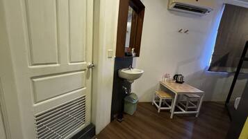 Uma casa de banho compacta com porta branca, lavatório com espelho, pequena mesa com cadeira e unidade de ar condicionado na parede.