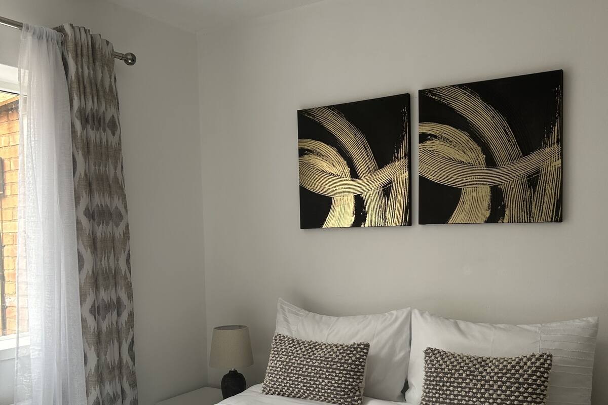 A neatly made bed with white linens and two pillows, a bedside lamp, and two abstract paintings on the wall.
