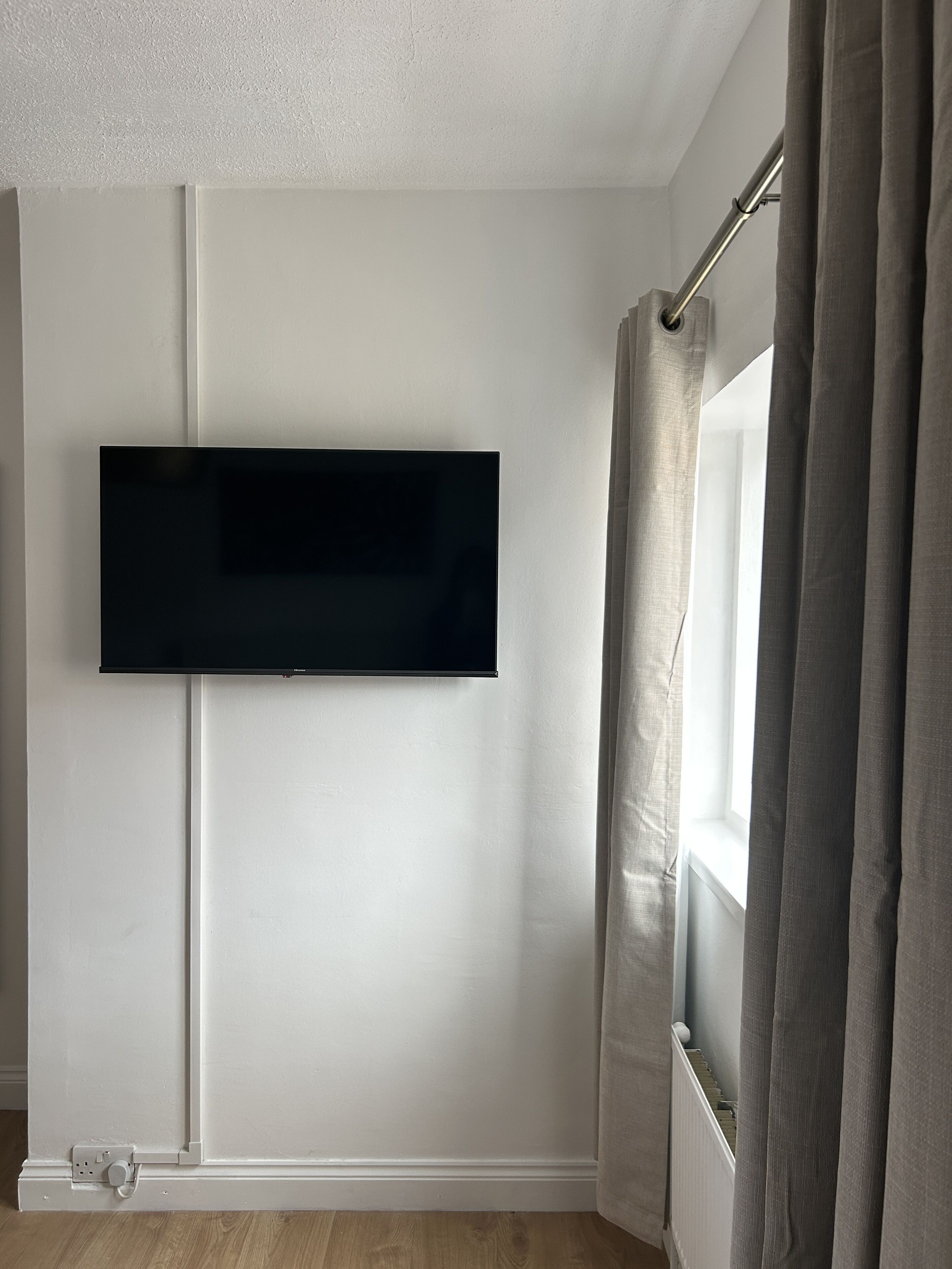 A room with a mounted flat-screen TV on the wall, a window with curtains, and a radiator below the window.