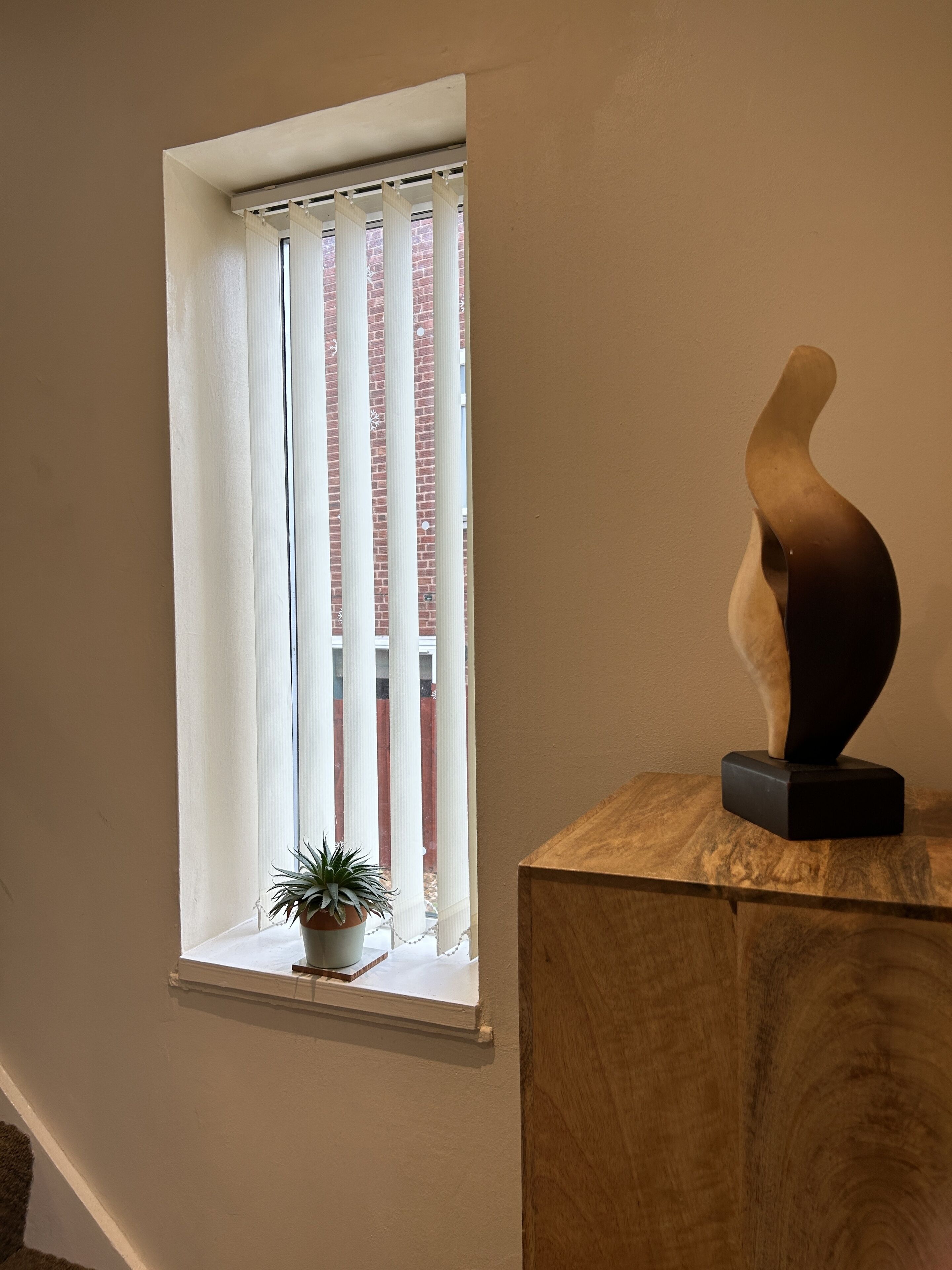 A window with vertical blinds, a potted plant on the sill, and a wooden sculpture on a pedestal.