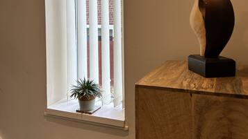 A window with vertical blinds, a potted plant on the sill, and a wooden sculpture on a pedestal.