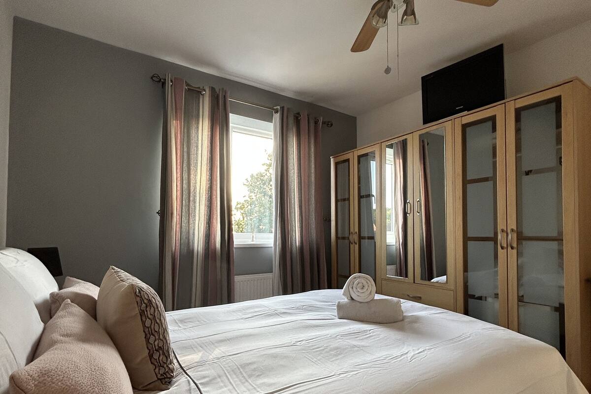 A bedroom with a large bed, a ceiling fan, a wardrobe, and a window with curtains.
