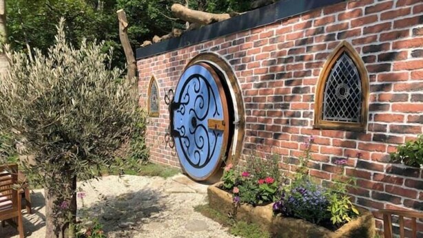 A brick building with a large circular window and a smaller arched window. There is a tree and flower beds in front of the building. - The Hobbit du Domaine de la Calotine (Dives-sur-Mer)