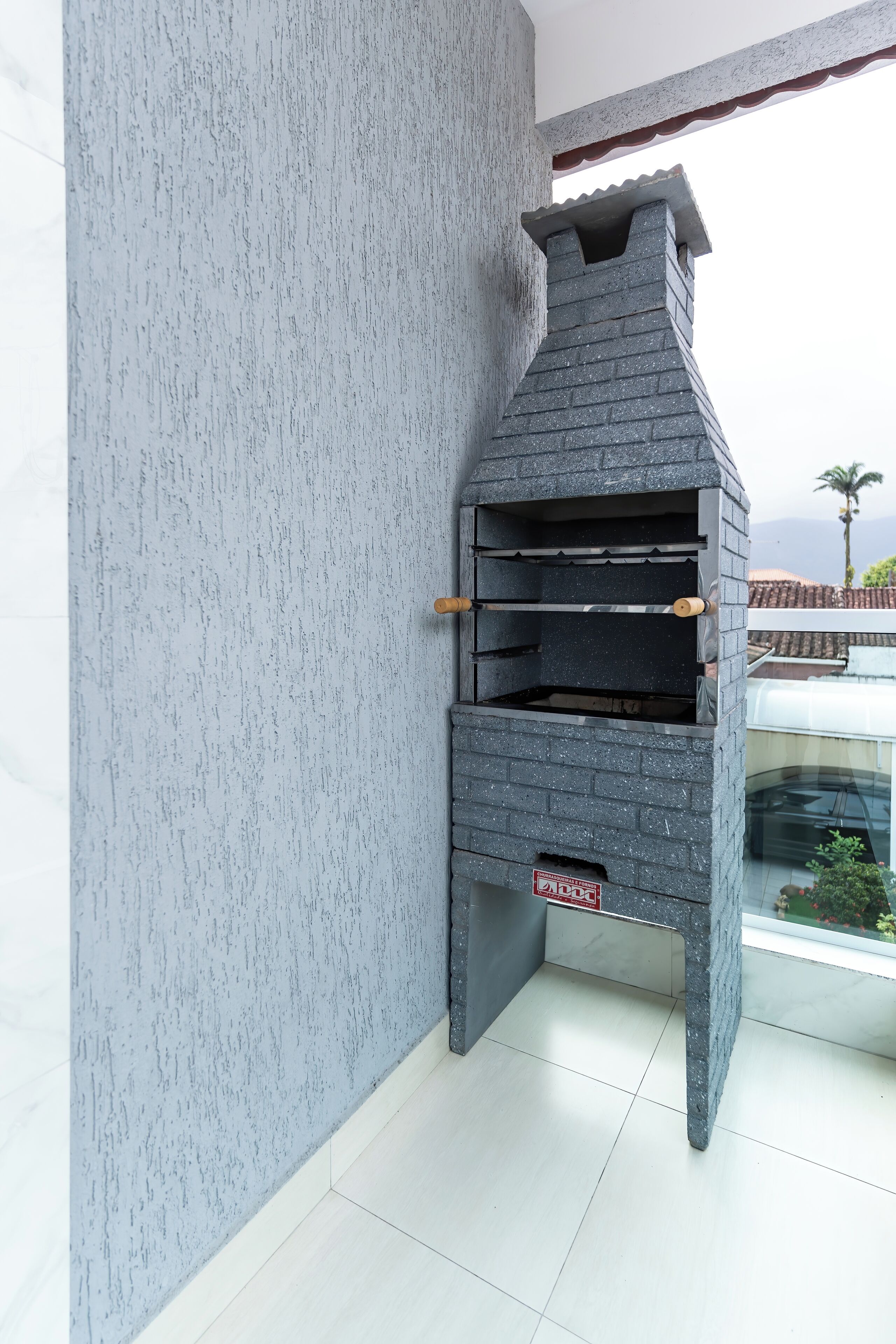 Forno in pietra all'aperto con canna fumaria, addossato a una parete strutturata, su un balcone con vista su una palma e dolci colline in lontananza.
