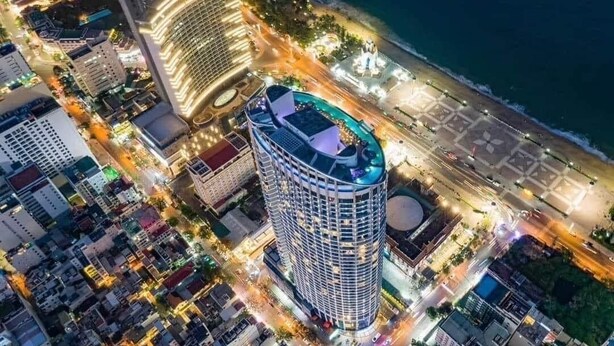 Ein Luftbild eines modernen Hotels mit bogenförmiger Fassade, gelegen in der Nähe eines Strandes und einer belebten Straße.
