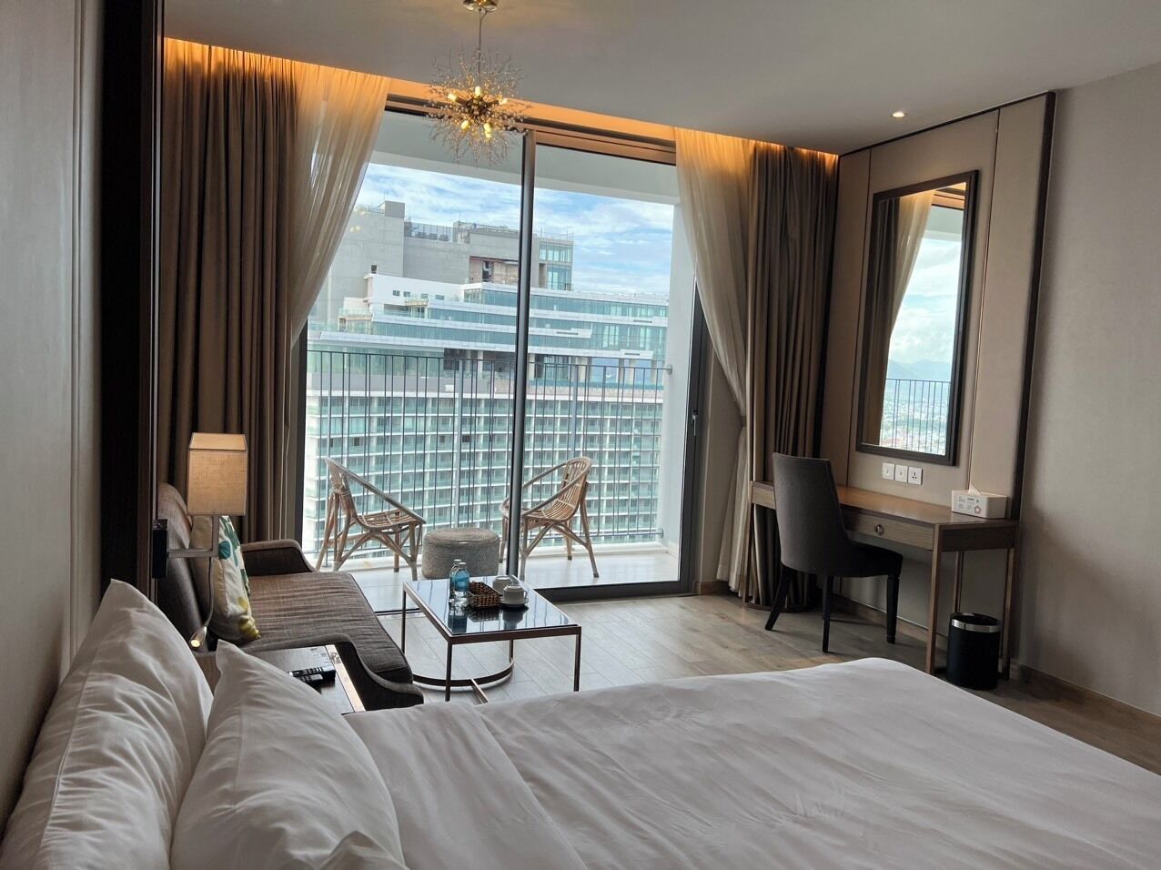 A hotel room with a large bed, a desk, a chair, a small table, and a balcony with chairs.