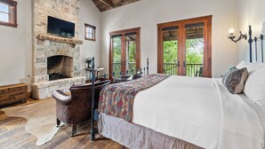Une chambre avec une cheminée en pierre, un lit recouvert d’une couverture à motifs, une chaise en bois et une vue sur la verdure par les fenêtres.
