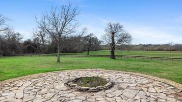 A stone-paved circular area with a fire pit in the center, surrounded by a grassy field and trees.