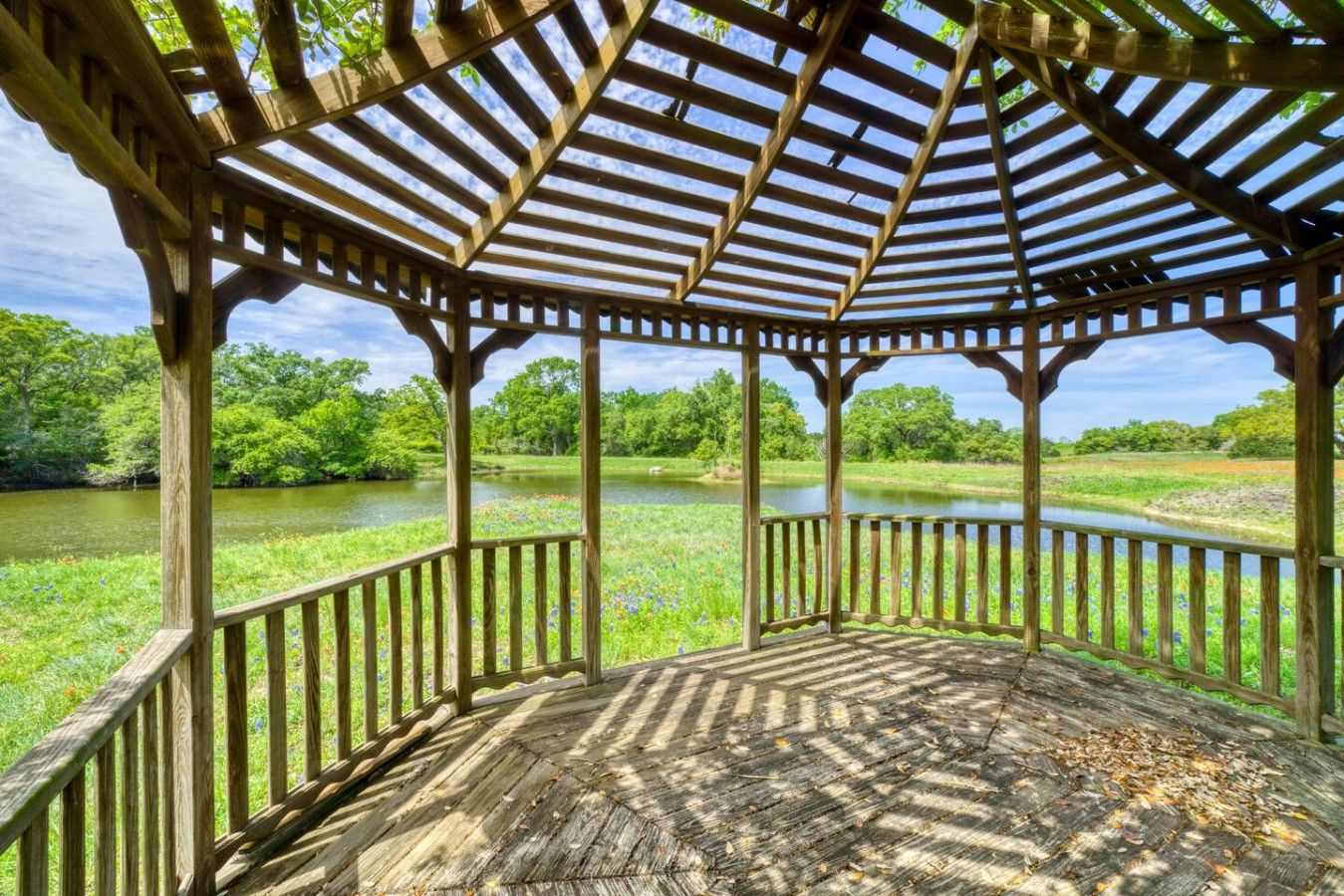 A wooden gazebo with a roof, surrounded by a natural landscape and a body of water.