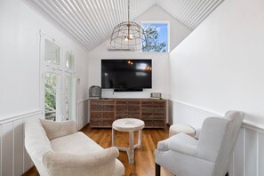 A room with a large flat-screen TV mounted on a wooden cabinet, two white armchairs, a small round table, and a ceiling light fixture. - Lakefront Charm: Your Perfect Getaway in Round Top (Fayette County)