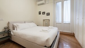 A neatly made bed with white linens, a bedside table with a lamp, and a radiator on the wall.
