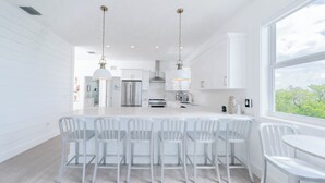 Uma cozinha moderna com armários brancos, uma ilha central com banquetas, iluminação pendente e uma vista através de uma grande janela.