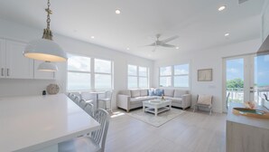 Uma sala de estar ampla com sofá branco, mesa de jantar com cadeiras e cozinha com armários brancos e luminária pendente.