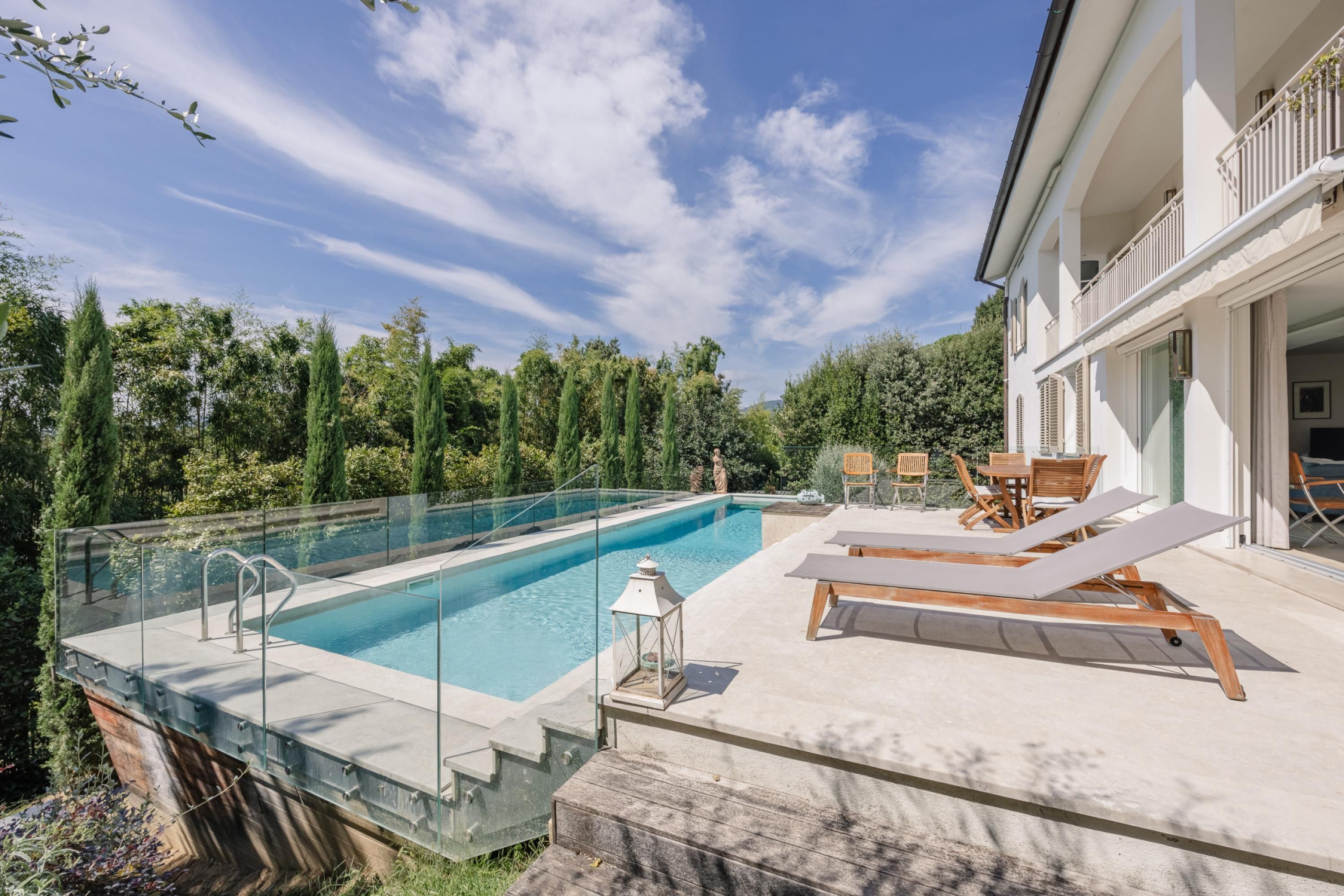 A modern outdoor swimming pool with clear glass walls, surrounded by lush greenery and a wooden deck with lounge chairs and a table.