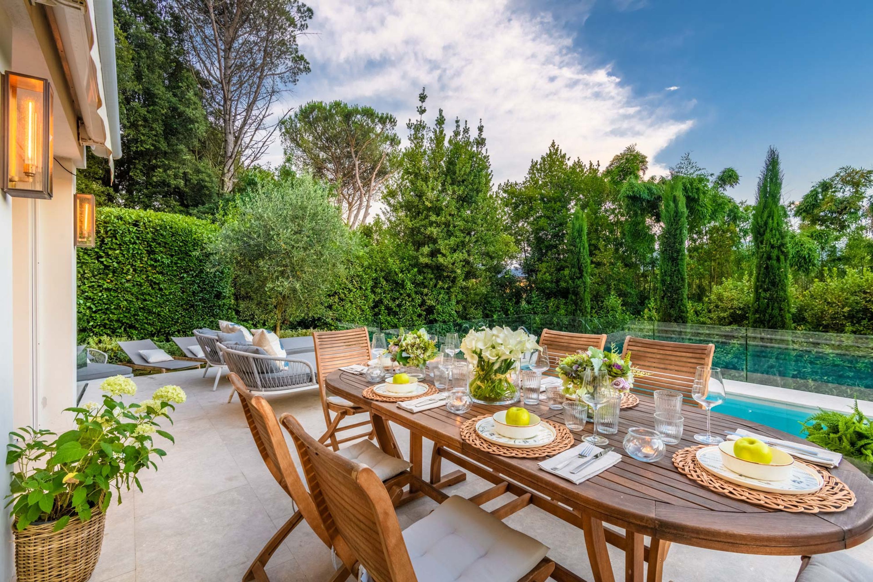 An outdoor dining area with a pool view, wooden table, chairs, and greenery.