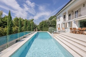 Una piscina rettangolare con acqua cristallina e blu, circondata da una barriera in vetro. Scale conducono a un pontile in legno con mobili da esterno.