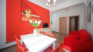 A dining area with a white table and orange chairs, a red accent wall with a painting, and a red sofa in the background.