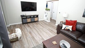 Ein Wohnzimmer mit einem dunkelbraunen Ledersofa, einem Holztisch, einem an der Wand montierten Fernseher, einem weißen Schrank und einem Korbwannenkorb am Boden.