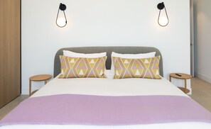 Une chambre à coucher moderne avec un grand lit, deux coussins à motifs, une tête de lit grise et deux tables de chevet avec des lampes.
