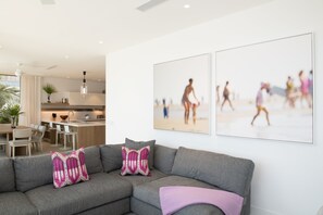 Un salon moderne avec un canapé d’angle gris, un coin repas avec une table et des chaises, et une cuisine en arrière-plan. Deux tableaux encadrés représentant des scènes de plage sont accrochés au mur.