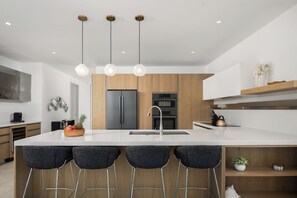 Une cuisine moderne dotée d’un grand îlot central, de tabourets de bar, de meubles en bois, d’appareils électroménagers en acier inoxydable et de luminaires suspendus.