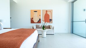 Una habitación de hotel moderna con una cama grande, un sofá, una mesa de centro con flores y dos cuadros abstractos en la pared.