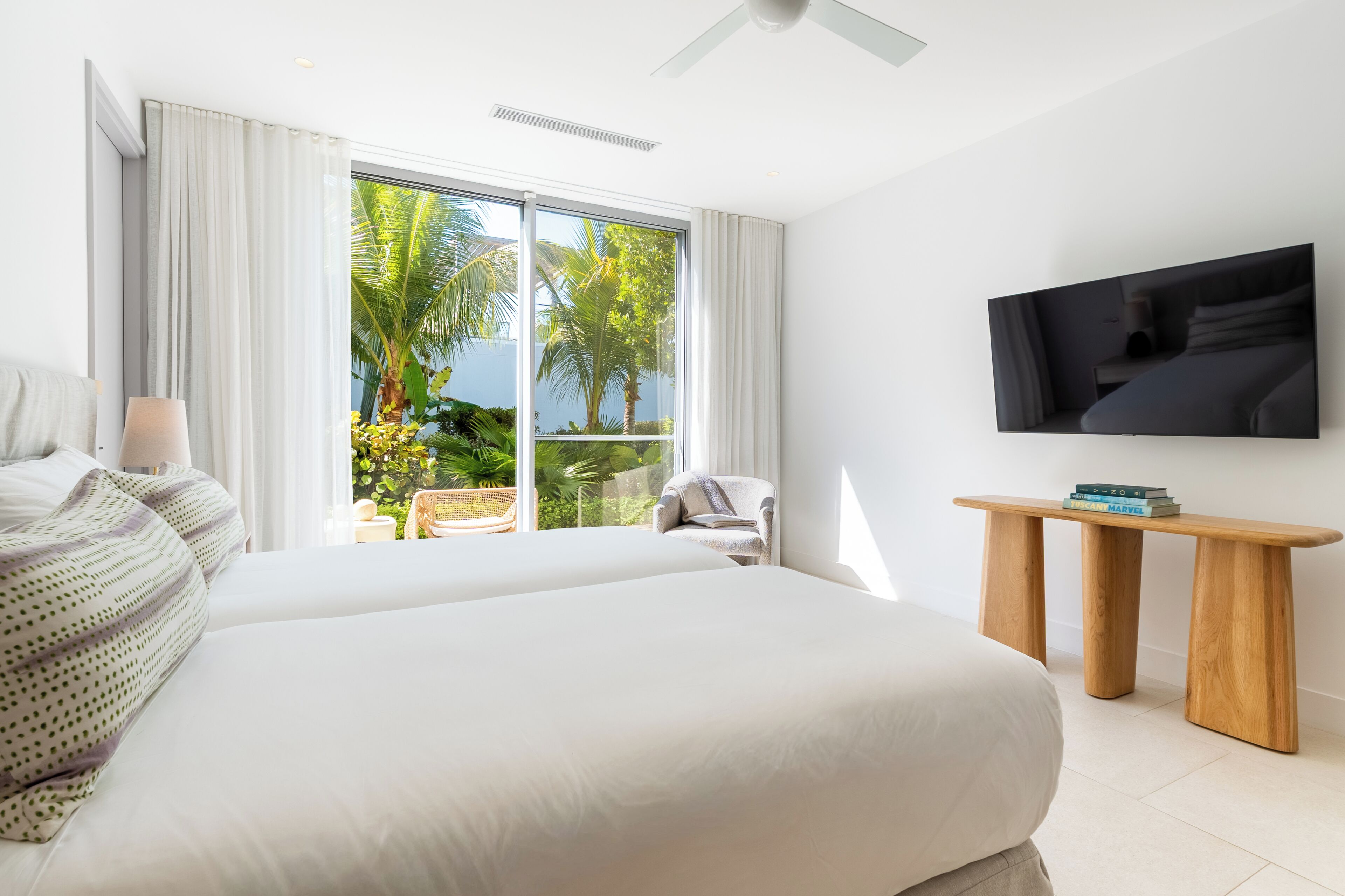 A hotel room with a large bed, a TV mounted on the wall, and a balcony with a view of palm trees.