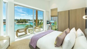 A modern bedroom with a large bed, a view of a beach and ocean, and a balcony with a chair and table.