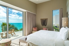 A bedroom with a large bed, a balcony with a view of the ocean, and a seating area with a table and chairs.