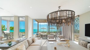 A modern living room with a large window overlooking a pool and ocean, a dining area with a table and chairs, and a comfortable seating area with sofas and a TV. - Ocean Estate Reef 5 Bedroom Including Guesthouse OE10 (Providenciales)