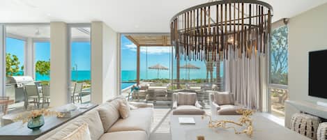 A modern living room with a large window overlooking a pool and ocean, a dining area with a table and chairs, and a comfortable seating area with sofas and a TV.