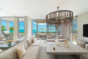 Un salon moderne doté d’une grande fenêtre donnant sur une piscine et l’océan, un coin repas avec une table et des chaises, et un espace salon confortable avec des canapés et un téléviseur.