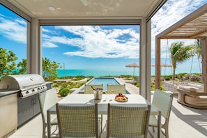 Un patio con una parrilla, mesa para comer y sillas, con vista a una piscina y al océano.
