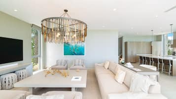 A modern living room with a large flat-screen TV, a dining area with a central table, and a kitchen with a bar.