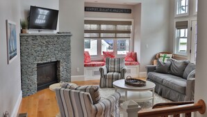 A living room with a stone fireplace, a flat-screen TV, a couch, striped armchairs, and a window with a view of the outside.