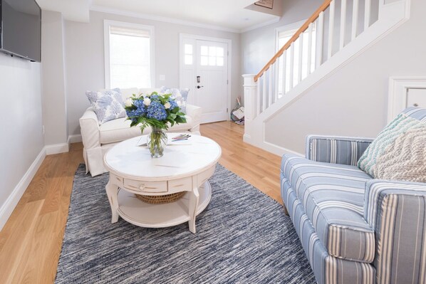 A living room with a white sofa, a striped couch, a round coffee table with a vase of flowers, and a staircase with white railings.