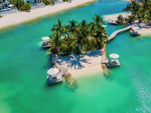 An aerial view of a tropical island resort with clear turquoise waters, palm trees, and a wooden dock leading to a lounge area with umbrellas.