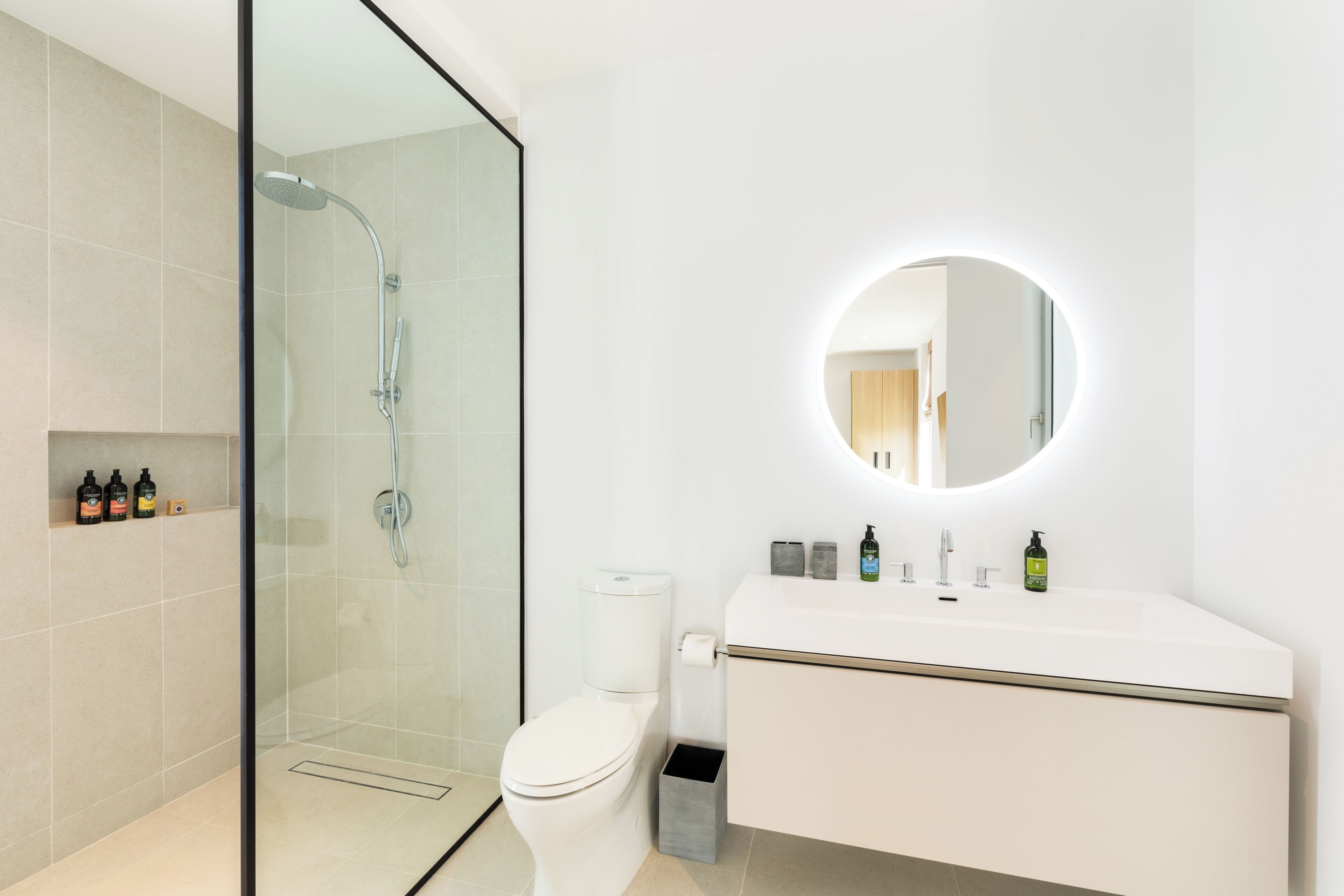 Une salle de bain moderne avec une douche à parois de verre, des toilettes blanches et un meuble-lavabo agrémenté d’un miroir rond.