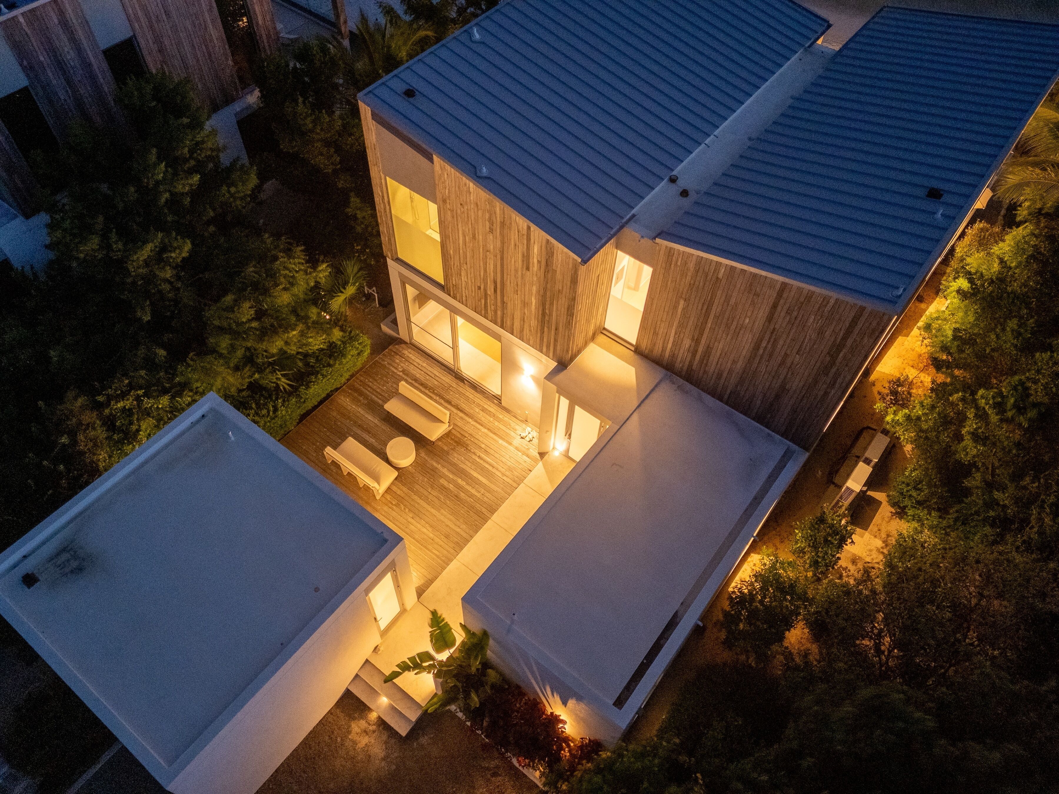 Une maison moderne à deux étages, dotée d’un toit plat, d’une terrasse et d’un petit espace extérieur aménagé pour s’asseoir.