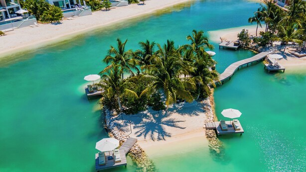 Vista aérea de un resort en una isla tropical con aguas turquesas cristalinas, palmeras y un muelle de madera que conduce a un área de lounge con sombrillas.